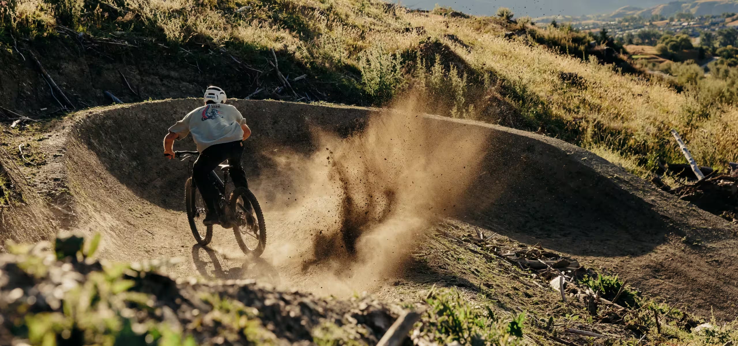 Person riding a bicycle on a dirt track with dust in the air, surrounded by natural landscape.